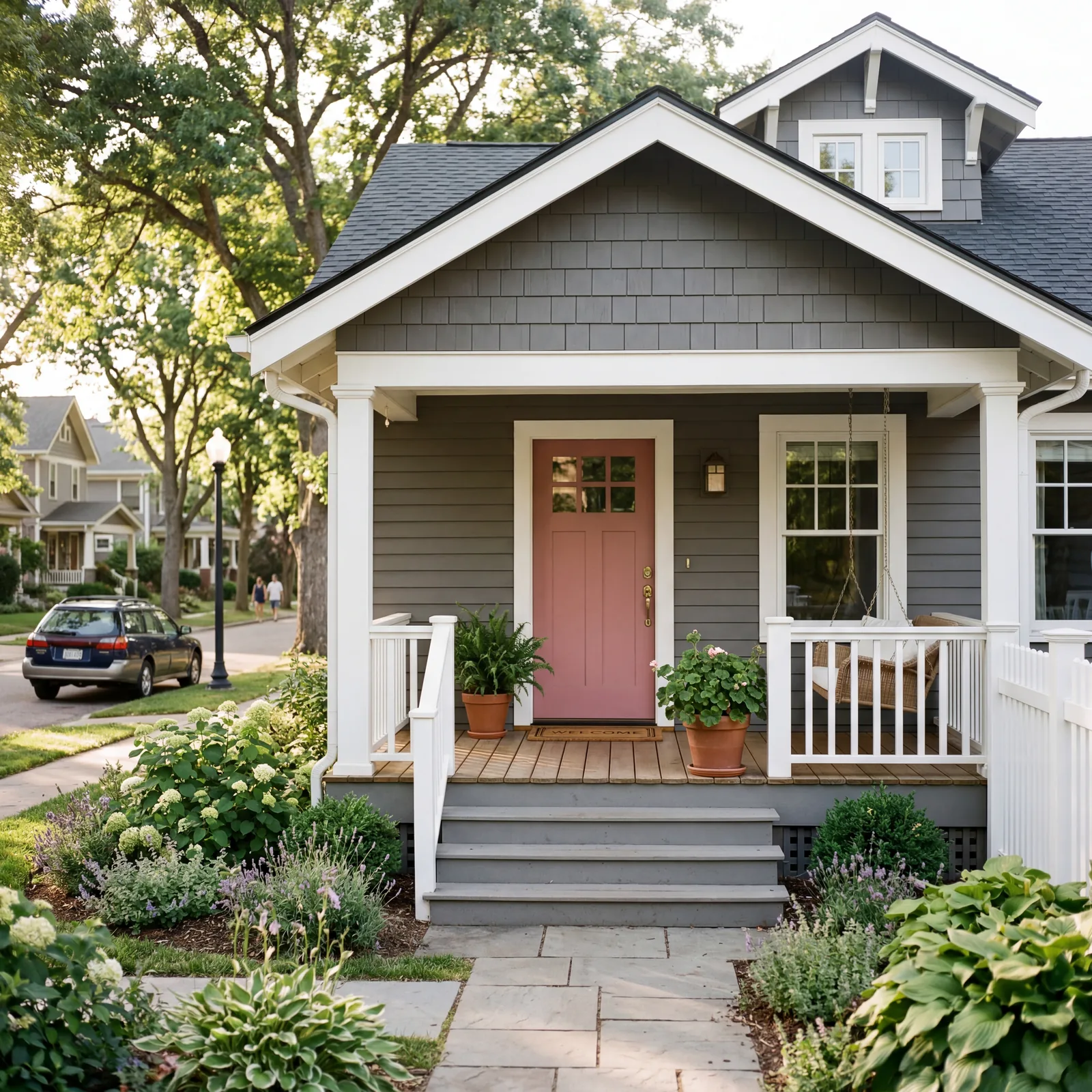 Grey craftsman bungalow with a dusty rose-pink painted front door, white porch railing, simple potted greenery, soft aft