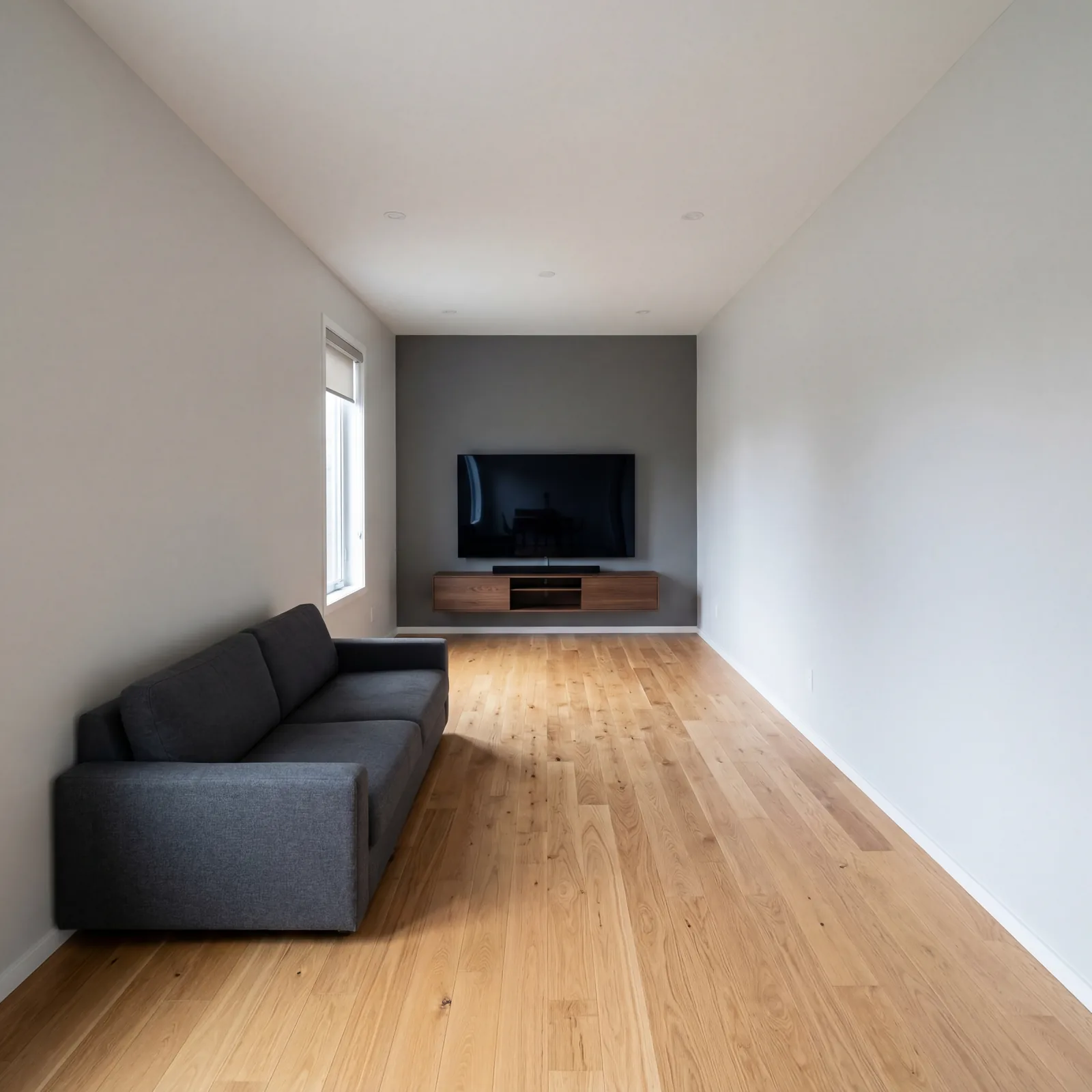 Living room with sofa pushed flat against the back wall facing a television on the opposite wall, with an empty expanse 