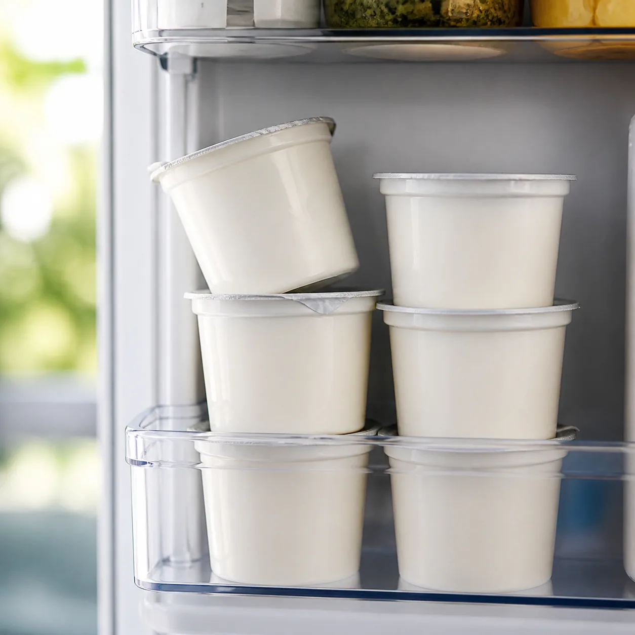 Realistic editorial photo of several yogurt cups stacked in a refrigerator door bin, one cup slightly tilted, soft dayli