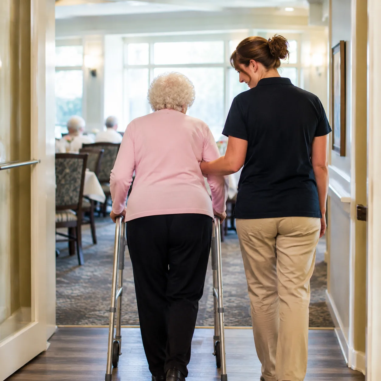 older resident using walker with caregiver escort toward assisted living dining room, realistic editorial lifestyle phot