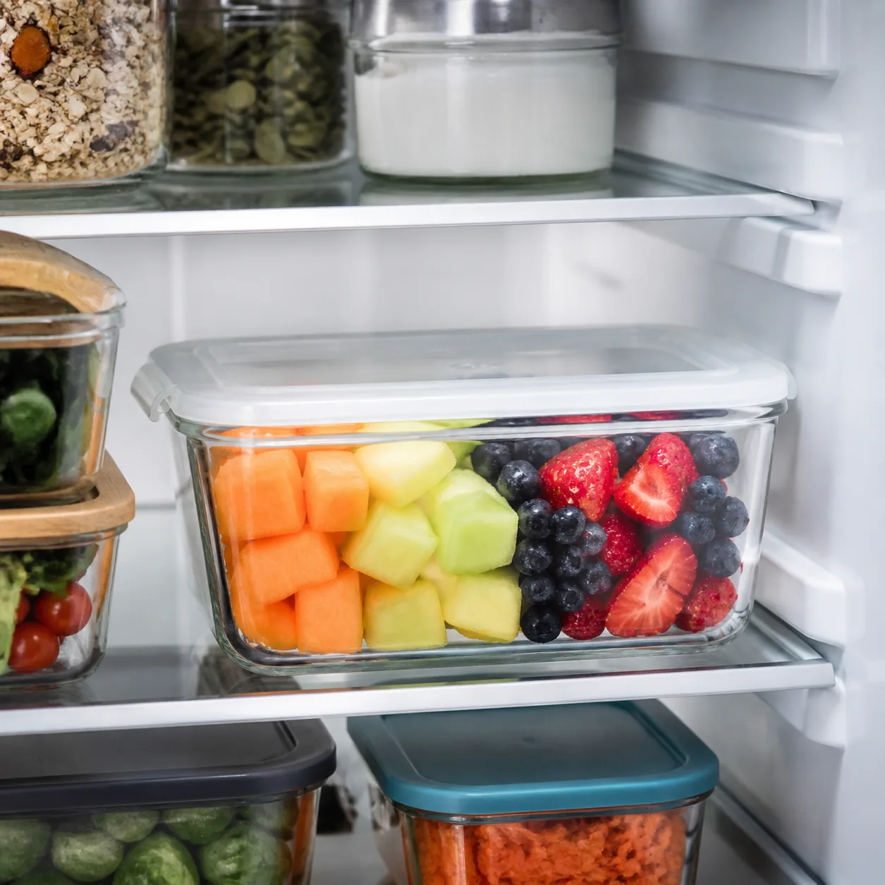 Realistic editorial photo of a container of cut melon and berries on a refrigerator shelf, clean meal prep containers, 1