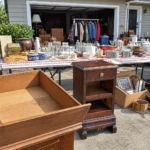 Realistic editorial photo of an American garage sale with folding tables, boxes, old kitchen items, furniture, and collectibles showing maker's marks under draw